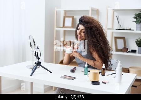 Happy cute gebräunt schöner lockiger lateinischer Beauty-Blogger in Casual Top erzählt über Gesichtscreme im Heim-Interieur. Space Mockup Banner kopieren. Einflussnehmer Stockfoto