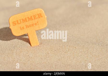 Minimaler Sommerhintergrund, orangefarbenes Holzschild im Strandsand an einem sonnigen Tag mit Text der Sommer ist da! Warmes Farbdesign mit Platz für Kopien Stockfoto