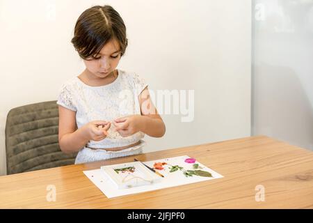 Kind mit weißem Papier, Blättern und Blumen, das Herbarium auf einem Esstisch aus Holz zu Hause macht. Getrocknete Blumenkunst Stockfoto
