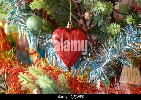 Nahaufnahme eines vollständig verzierten Weihnachtsbaums mit einer markanten roten herzförmigen Kugel, umgeben von buntem Lametta. Stockfoto