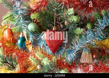 Nahaufnahme eines vollständig verzierten Weihnachtsbaums mit einer markanten roten herzförmigen Kugel, umgeben von buntem Lametta. Stockfoto