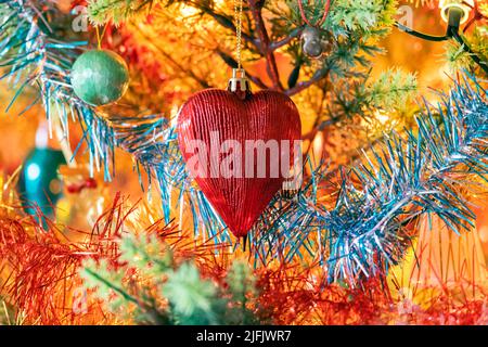 Nahaufnahme eines vollständig verzierten Weihnachtsbaums mit einer markanten roten herzförmigen Kugel, umgeben von buntem Lametta. Stockfoto