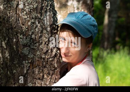 Junge Frau in pinkem Hemd und Jeans-Mütze, Kopfhörer umarmt eine Birke und lächelt Stockfoto