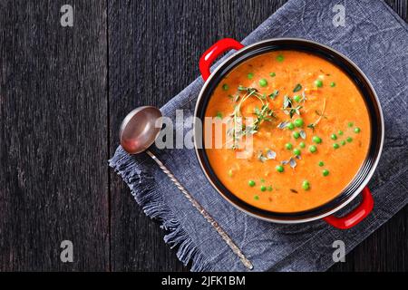 Gebratene, dicke Yamssuppe mit Gemüse in rotem Topf auf einem dunklen Holztisch mit Suppenkelle, horizontale Ansicht von oben, flach liegend, freier Platz Stockfoto