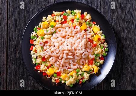 Gekochte kleine rosafarbene Garnelen auf Hirsepilaff mit Gemüse, Mangostückchen und Gewürzen auf schwarzem Teller auf dunklem Tisch, horizontale Ansicht von oben Stockfoto