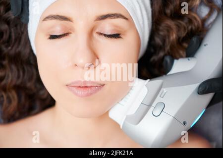 Nahaufnahme Porträt von jungen schönen Frau mit lockigen Haaren tun Hautpflege Verfahren. Konzept des Prozessablaufs mit moderner Ausstattung in der professionellen Klinik. Stockfoto