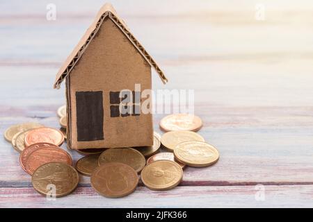 Papierhaus Spielzeug oder Papphaus und Münzen auf Holztisch mit Morgensonne. Kopieren Sie Platz für Design und Nutzung. Immobilien- und Finanzkonzept. Stockfoto