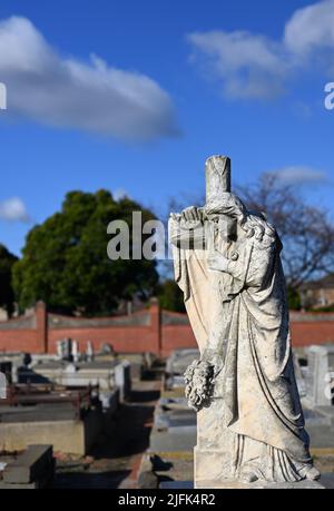 Abgenutzte Steinskulptur einer Frau, die trauert, während sie sich auf ein christliches Kreuz oder Kruzifix stützt, mit Grabereihen im Hintergrund Stockfoto