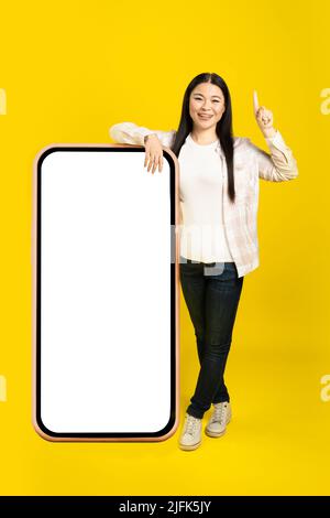 Mit dem Finger nach oben gelehnt lehnte sich die asiatische Frau des mittleren Alters auf ein riesiges Smartphone mit weißem, blanken Bildschirm und trug ein lässiges Outfit, das auf gelbem Hintergrund isoliert war. Modell mit freiem Speicherplatz. Stockfoto