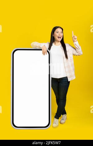 Asiatische Frau mittleren Alters aufgeregt Zeigefinger nach oben gelehnt auf riesige, riesige Smartphone mit weißen weißen Bildschirm, tragen lässige Outfit isoliert auf gelbem Hintergrund. Modell mit freiem Speicherplatz. Stockfoto