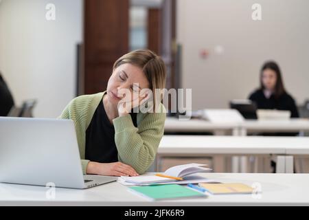 Erschöpfte reife Schülerin schläft während des Online-Unterrichts ein und lernt im Klassenzimmer auf einem Laptop Stockfoto