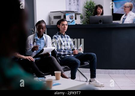 afroamerikanischer Arzt, der mit Patienten an der Krankenhausaufnahme spricht und Termine und Prioritäten der Patienten überprüft. Asiatischer Patient wartet in der Privatklinik, während der Arzt die Kliniktriage macht. Stockfoto