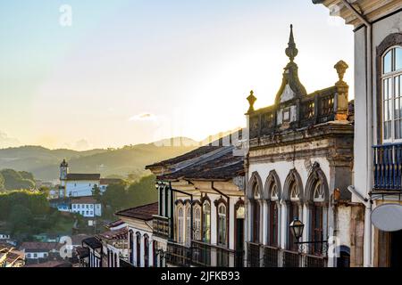 Blick auf koloniale Häuser Fassade und historische Barockkirche im Hintergrund auf den Hügeln von Ouro Preto Stadt, Minas Gerais Staat, Brasilien.