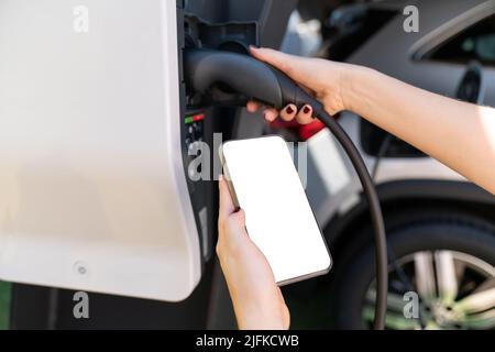 Hände mit Smartphone und Ladestecker auf einem Hintergrund von Elektroauto . Weißer Bildschirm, können Sie hier Ihre Inhalte hinzufügen Stockfoto