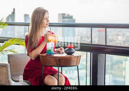 Schöne junge Frau mit Cocktails ruht im luxuriösen Dachrestaurant. Elegante Dame in rotem Kleid mit Mocktails auf der Sky Bar Terrasse Stockfoto