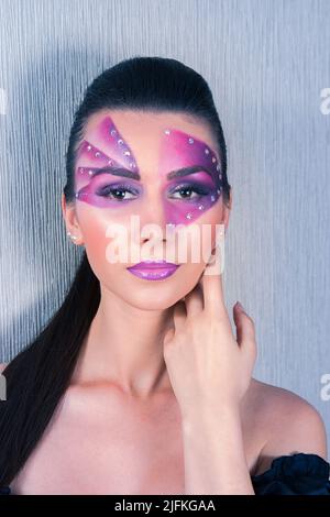 Wunderschöne kaukasische Frau mit violettem Gesichtsbemalung und Kristallen auf ihrem Gesicht, die ihre Wange und ihren Hals berühren, auf einem silbernen Hintergrund Stockfoto