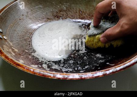 Eine verbrannte, verbrannte und fettige Pfanne oder einen Wok auf dem Spülbecken mit menschlicher Hand putzen oder reinigen. Stockfoto