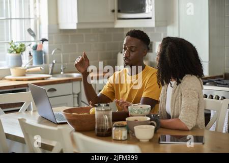 Junges, multiethnisches Paar, das beim Frühstück mit einem Laptop spricht Stockfoto