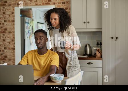 Ein junges, multiethnisches Paar, das morgens in der Küche einen Laptop benutzt Stockfoto