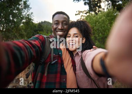 Ein junges, multiethnisches Paar lächelt und macht Selfies auf einem Wanderweg Stockfoto