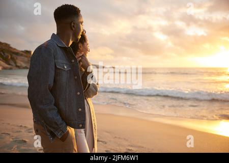 Junges multiethnisches Paar, das an einem Strand spazieren geht und den Sonnenuntergang betrachtet Stockfoto