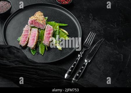 Hausgemachtes geschnittenes gegrilltes Sesam-Thunfischsteak mit Frühlingszwiebeln und Zuckerschoten, auf Teller, auf schwarzem Steinhintergrund, mit Copyspace und Platz für Stockfoto
