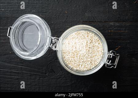 Gesundes Lebensmittelkonzept. Sesamsamen, im Glasglas, auf schwarzem Holzgrund, von oben flach liegend Stockfoto