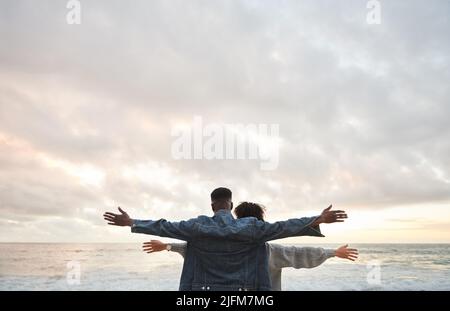 Junges multiethnisches Paar, das mit erhobenen Armen am Strand steht Stockfoto