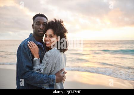 Junges multiethnisches Paar, das Arm in Arm am Strand bei Sonnenuntergang steht Stockfoto