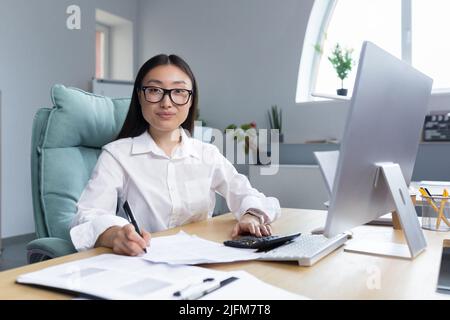 Porträt einer jungen, schönen und erfolgreichen asiatischen Geschäftsfrau, lächelnder weiblicher Angestellter und Blick auf die Kamera, Finanzbuchhalter Papierkram. Stockfoto