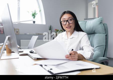 Porträt eines jungen erfolgreichen asiatischen Finanziers, einer Geschäftsfrau, die mit Dokumenten im Büro arbeitet, lächelt und die Kamera anschaut. Stockfoto