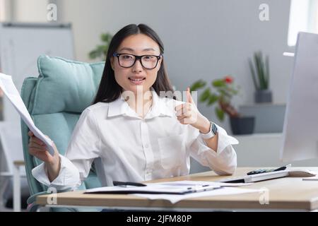 Junge Finanzfrau, die lächelt und die Kamera anschaut, asiatische Geschäftsfrau, die die Kamera anschaut und lächelt, hält einen guten Finanzbericht in den Händen und zeigt den Daumen nach oben. Stockfoto