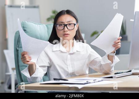 Frustriert und verärgert asiatische Geschäftsfrau, die im Büro arbeitet, Dokumente mit schlechten und negativen Finanzkennzahlen des Unternehmens hält, Finanzier arbeitet. Stockfoto