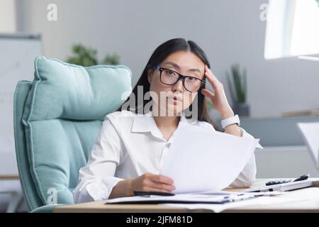 Frustriert und verärgert asiatische Geschäftsfrau, die im Büro arbeitet, Dokumente mit schlechten und negativen Finanzkennzahlen des Unternehmens hält, Finanzier arbeitet. Stockfoto