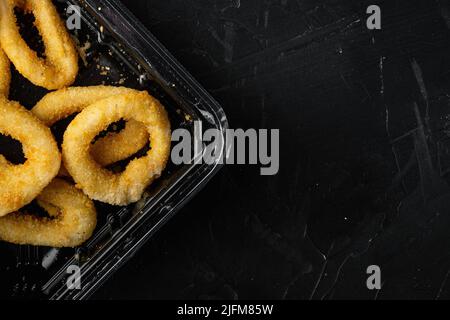 Paket mit frittierten Tintenfisch- oder Zwiebelringen, auf schwarzem dunklen Steintischhintergrund, flache Ansicht von oben, mit Platz für Text Stockfoto