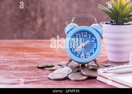 Leeres Notizbuch, Münzen, Wecker und Blumen auf dem Schreibtisch mit Holzhintergrund. Speicherplatz zur Verwendung kopieren. Geschäfts- und Bildungskonzept. Stockfoto