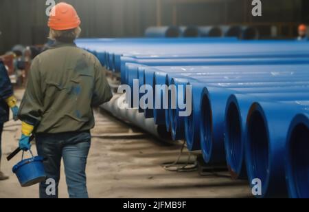 Arbeiterin in der Helmrückansicht in der Werkstatt mit großen Metallwasserrohren, die zum Lackieren bereit sind. Eisenrohrproduktion, Schwerindustrie. Stockfoto