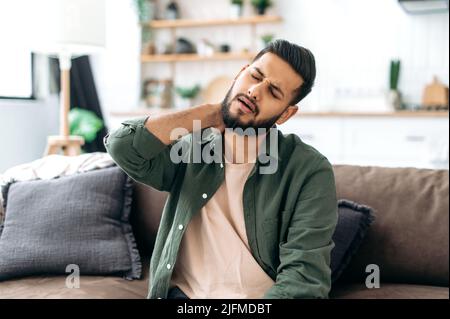 Verärgert indischen oder arabischen jungen Mann, sitzen auf dem Sofa in gemütlichen Wohnzimmer, massieren seinen Hals, müde nach einem harten Tag bei der Arbeit, leiden an Gelenkproblemen, eingeklemmt Nerven im Hals, brauchen Ruhe Stockfoto