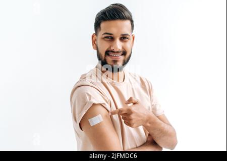 Impfkonzept. Junger inder oder araber, der einen Impfschutz erhält das Coronavirus und andere Viren, nach der Impfung Arm mit Gips zeigend, auf isoliertem weißen Hintergrund stehend, lächeln Stockfoto