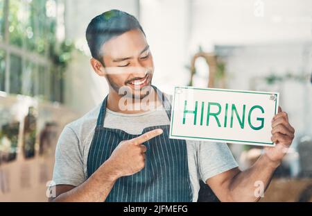 Ein junger hispanischer Mann zeigt auf ein Einstellungsschild an einem Fenster, das in einem Café oder Geschäft ausgestellt ist. Glücklich gemischt Rennen Kerl Werbung Job Gelegenheit, während Stockfoto
