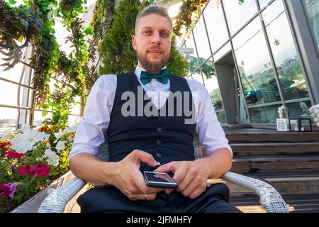 Ein junger, selbstbewusster Mann in einem weißen Hemd, einer schwarzen Weste mit einem Smartphone in den Händen schaut auf die Kamera, während er im Penthouse sitzt Stockfoto