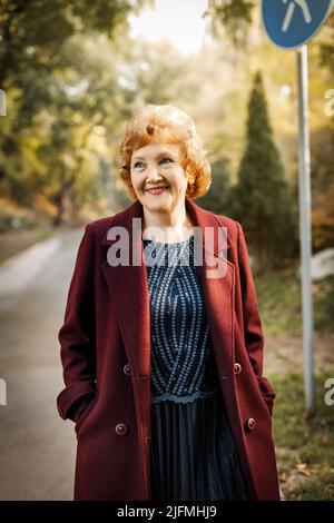Russische reife Frau mit kurzen Haaren lächelnd sieht im Herbst im Freien in einem nicht geknöpften burgunderfarbenen Mantel zur Seite Stockfoto