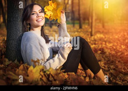 Junge glückliche zentralasiatische Frau, die in der Nähe von Baum sitzt und im Herbstpark den Sonnenuntergang anschaut Stockfoto