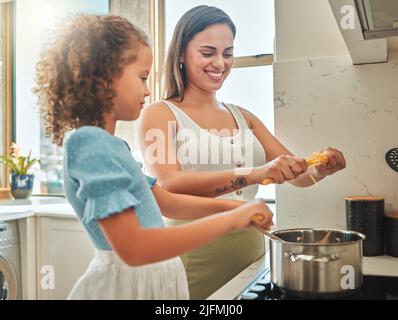 Mutter und kleine Tochter kochen gemeinsam in der Küche. Mutter und Kind aus gemischter Rasse stehen am Herd, brechen Spaghetti und werfen sie hinein Stockfoto