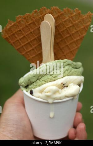 Eine Hand, die einen schmelzenden Matcha und eine Eisbecher mit Vanilleschokolade-Chips und einer Waffel hält Stockfoto