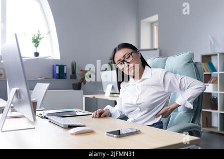 Müde weibliche Büroangestellte massiert ihren Rücken, asiatische Geschäftsfrau hat schwere Rücken- und Rückenschmerzen. Stockfoto
