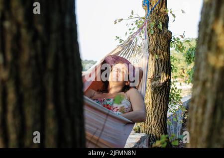 Junge Frau, die im Sommer in der Hängematte zwischen Bäumen liegt und sich bräunt. Stockfoto