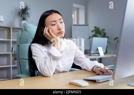 Eine traurige Geschäftsfrau arbeitet in einem modernen Büro, eine asiatische Frau denkt über die Ergebnisse der Arbeit nach. Stockfoto