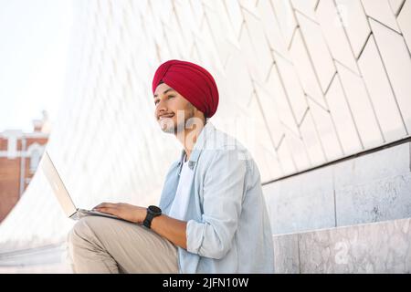 Optimistischer indischer Student, Angestellter, Freiberufler, der im Freien sitzt und einen Laptop benutzt, erfolgreicher junger hinduistischer Unternehmer in traditionellem Turban-Kopftuch mit Laptop, der auf der Treppe im Stadtbild sitzt Stockfoto
