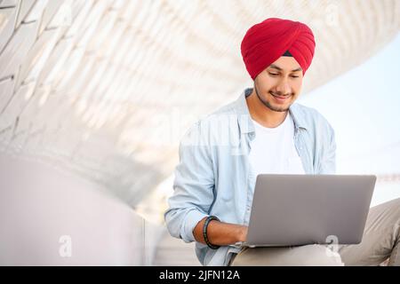 Indischer Freiberufler im traditionellen Turban Pagg sitzt im Freien mit Laptop, Entwickler arbeitet auf der Ferne, hindu Kerl genießt Remote-Arbeit, männliche Student studieren online, E-Learning-Konzept Stockfoto
